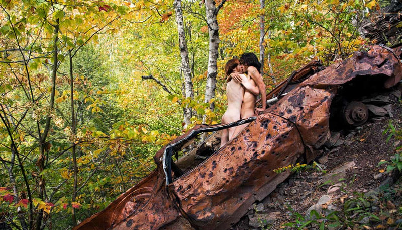 a gay couple kisses on top of a rusty car in the middle of the forest 