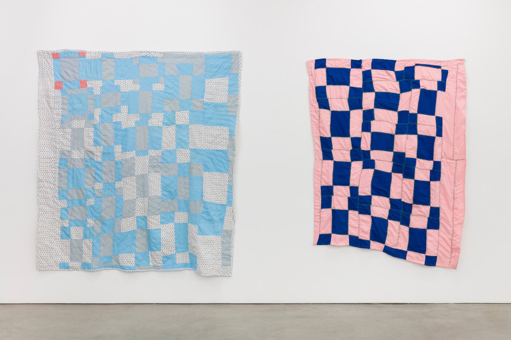 An installation view of two quilts by Gracie Scott. The quilt to the left is rendered in grey and blue squares with four small red squares at the top left corner. The right quilt is rendered in navy blue and light pink squares. 
