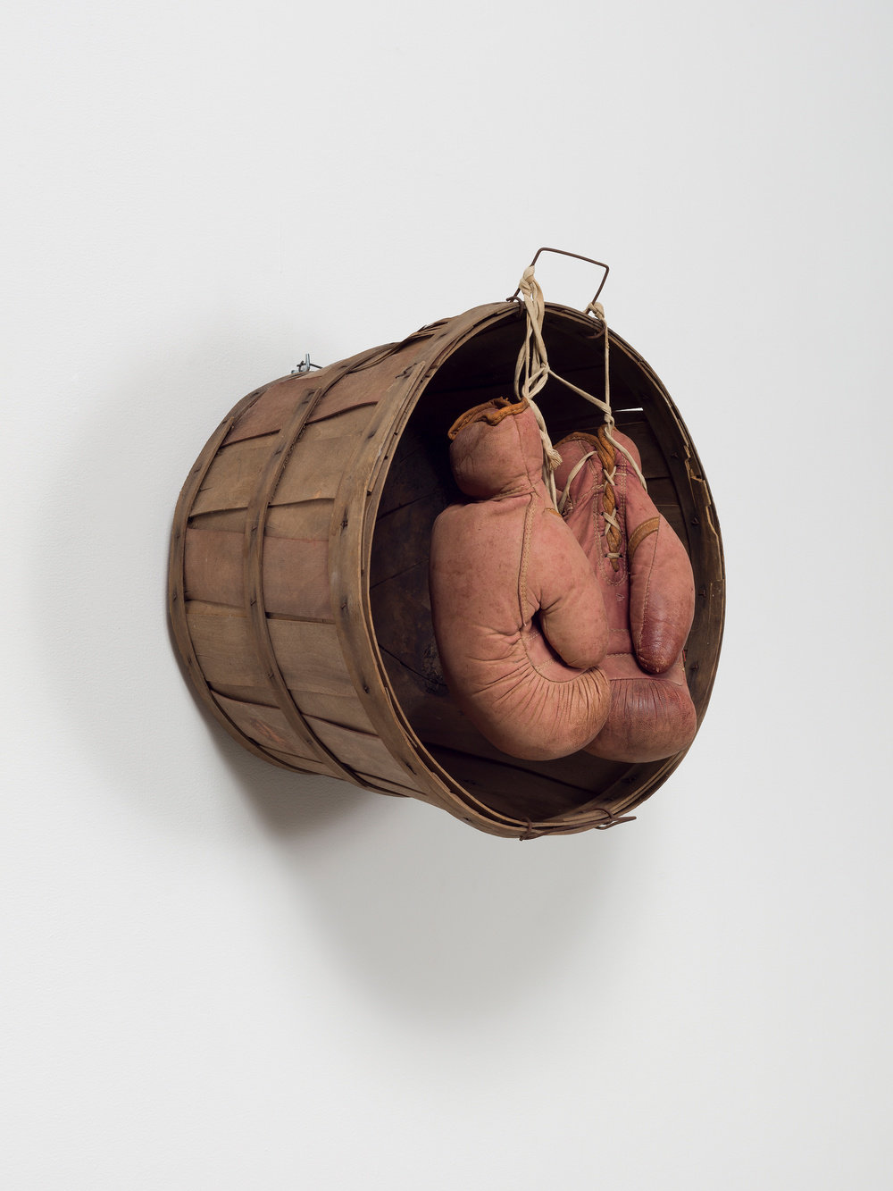 A sculpture by Lonnie Holley composed of worn boxing gloves hung from the handle of a wicker peach basket. The base of the basket is mounted to the wall.