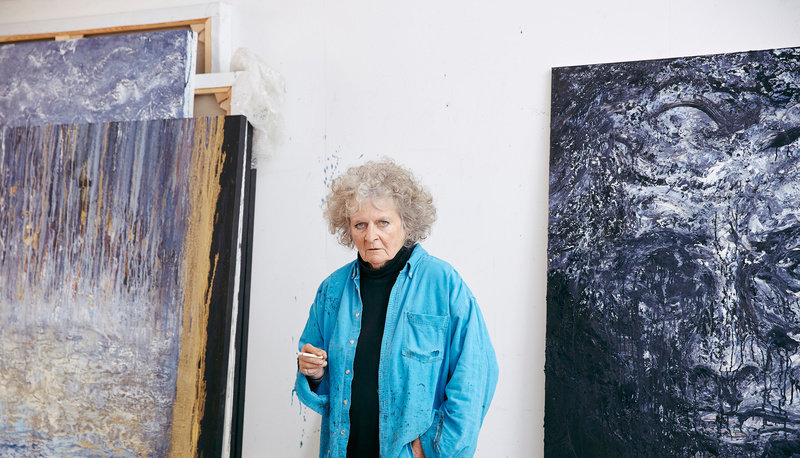 Maggi Hambling standing in front of a stack of her large paintings leaning against the walls beside her 